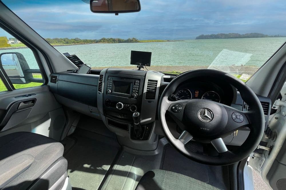 Motorhome cab interior with a Mercedes steering wheel, dashboard screen and estuary view through the windscreen