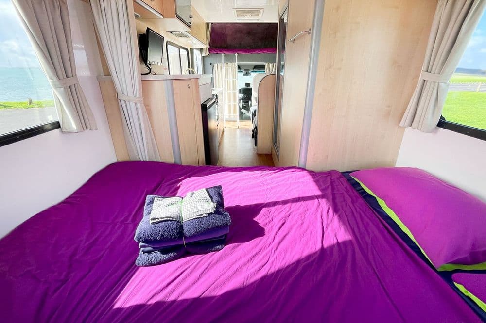 Rear double bed of the motorhome made up with purple bedding, windows on three sides and a corridor leading forward