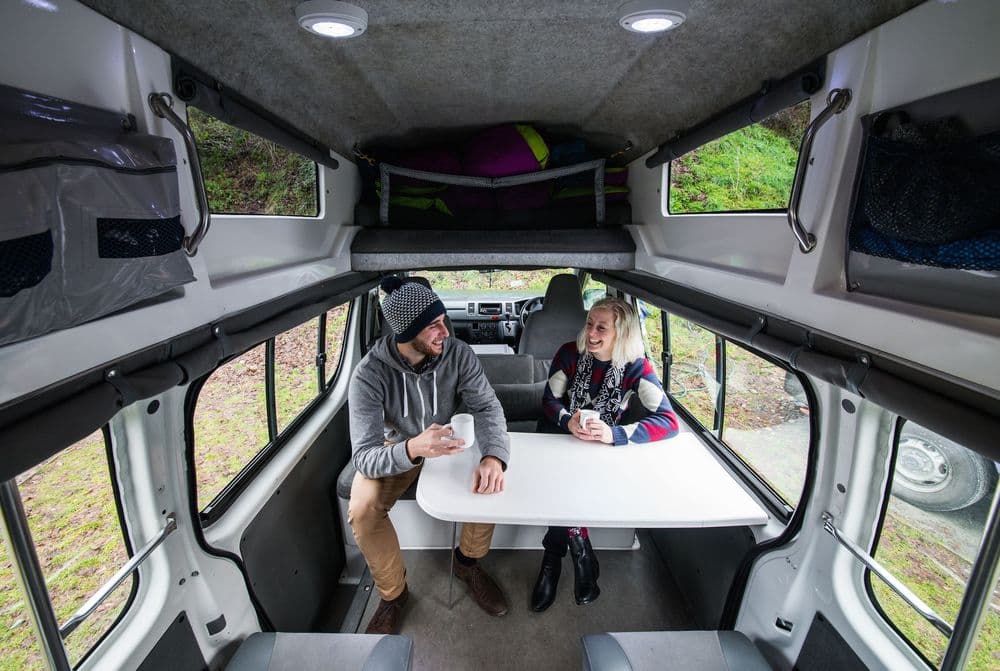 Interior of the campervan looking forward from the rear, with two people at the table and the roof bed made up above