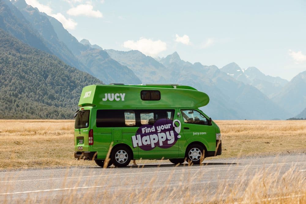 Green JUCY Toyota HiAce campervan with a large white over-cab sleeping pod, parked on a tussock flat below mountains