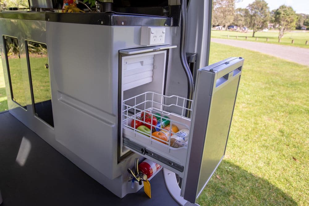Rear kitchen of the campervan with the fridge door open and a cutlery basket on the inside of the door