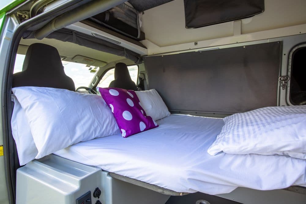 Two campervan beds made up with white linen and purple pillows, one in the roof pod and one below