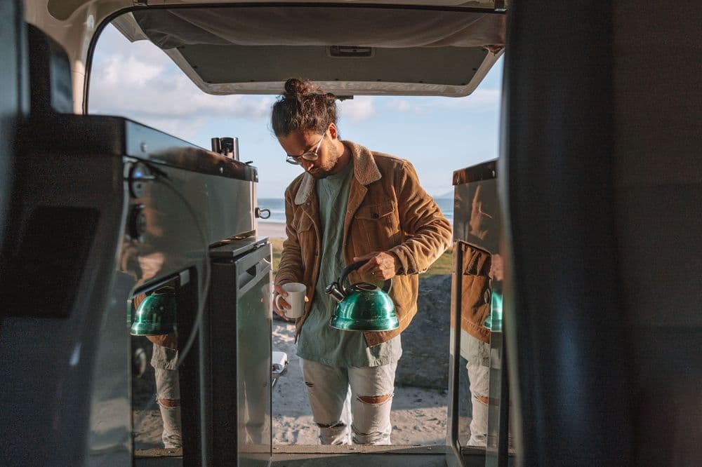 Person leaning into the rear of the campervan holding a kettle, sea and headland visible beyond the open doors