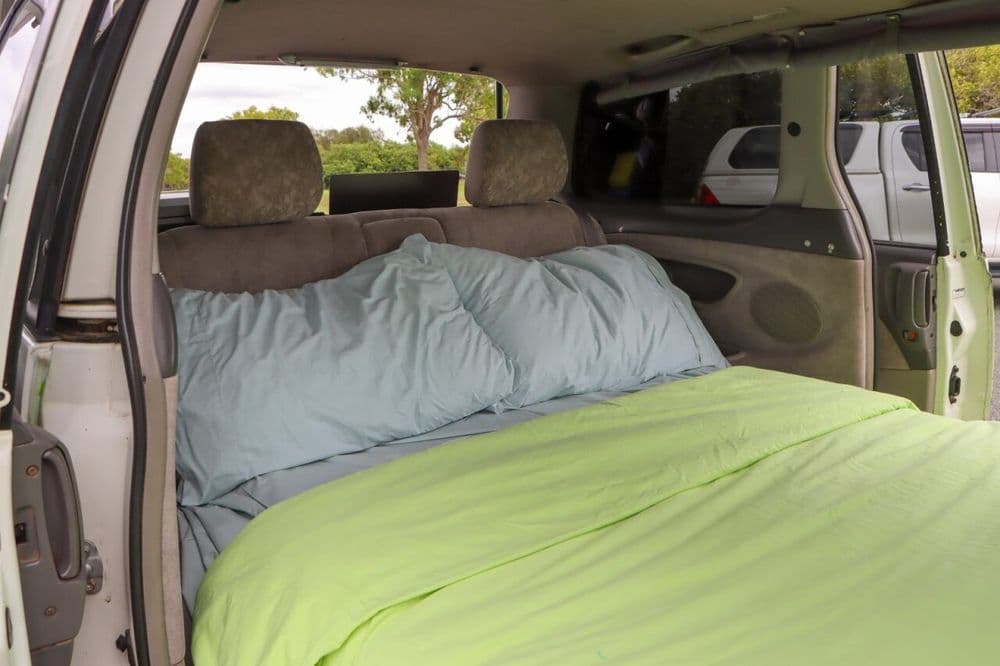 Interior of the campervan through the open side door, showing the made bed with green and grey bedding against the cabin trim