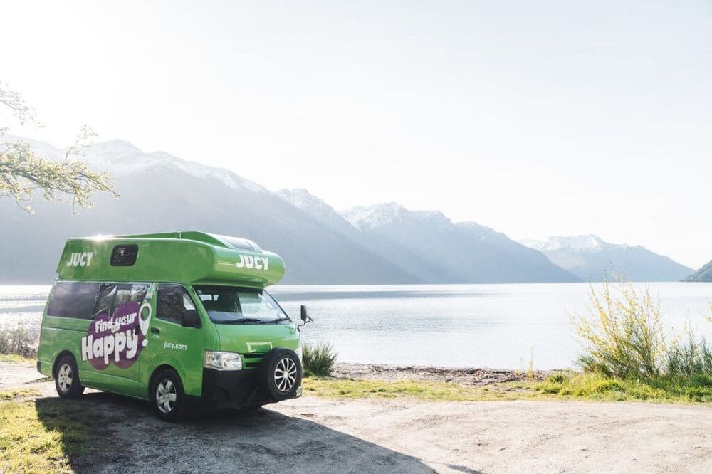 Green JUCY high-roof Toyota HiAce campervan with a raised roof pod parked on gravel beside a lake and mountains