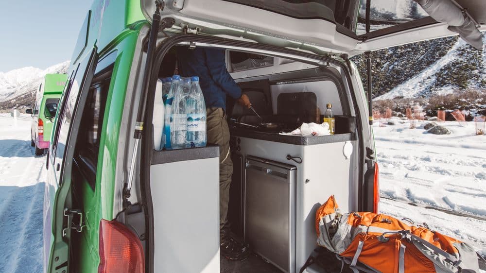 Rear doors of the green campervan open in a snowy car park, showing the kitchen unit and stored gear