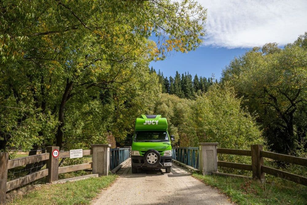 Green JUCY campervan driving across a single-lane timber bridge on an unsealed rural road between fences