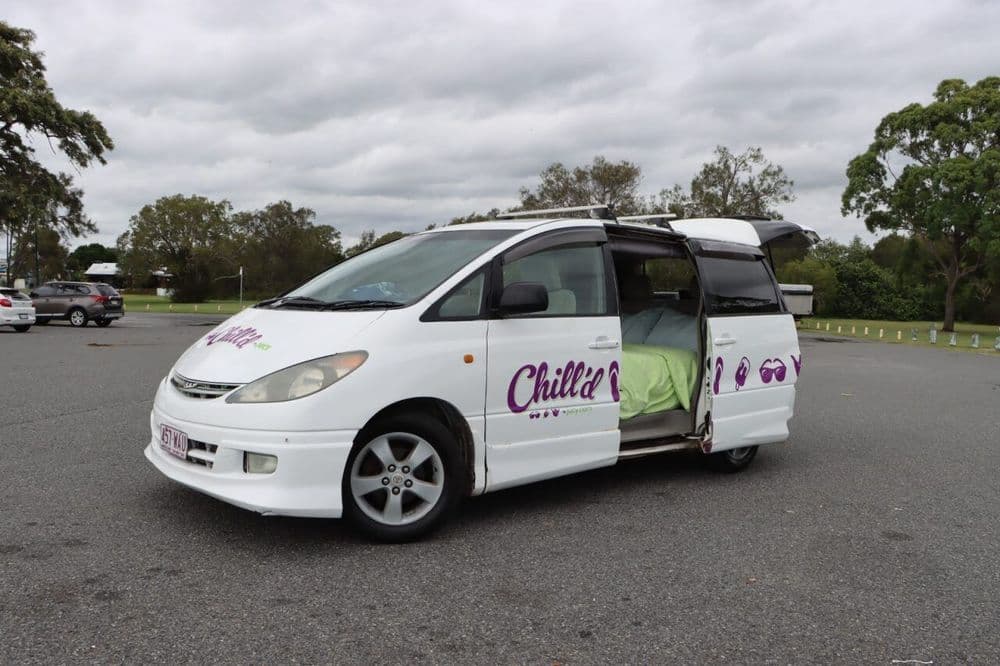 Side view of the white campervan on a car park by the sea with its side door open and the bed made up inside