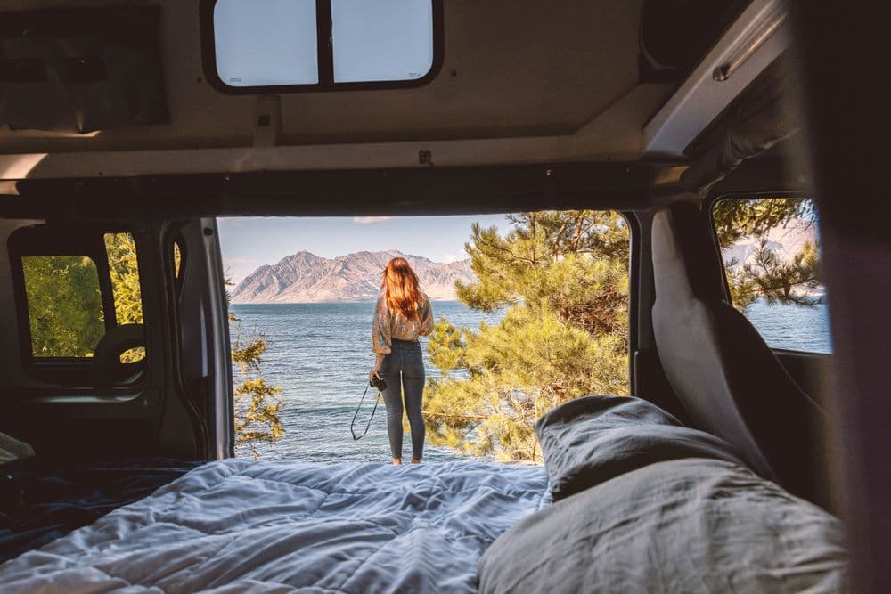 Lower double bed made up inside the campervan with the side door open onto a lake and a person standing outside
