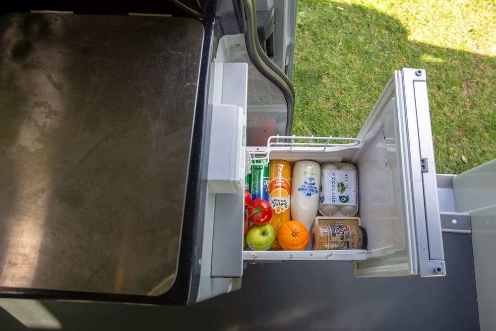 Open upright compressor fridge inside the campervan, stocked with fruit, vegetables, milk and bottles