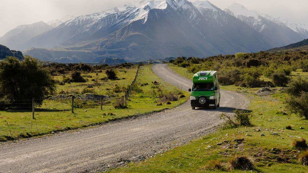 Green campervan driving away along a wide gravel road towards a snow-streaked mountain range
