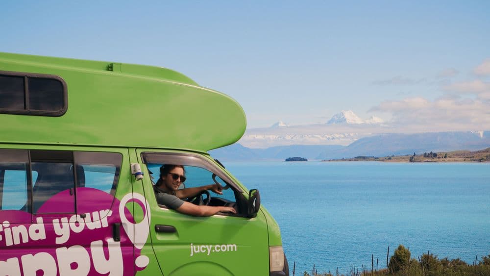 Driver at the wheel of the campervan smiling, with a lake and mountains through the side window